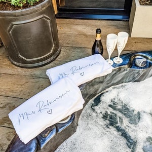 May include: Two white towels with the text "Mr. Robinson" and "Mrs. Robinson" with a heart symbol on each towel. The towels are placed on the edge of a hot tub with bubbles in the water. Two champagne flutes with the text "Mr. Robinson" and "Mrs. Robinson" are on the edge of the hot tub. A bottle of champagne is behind the flutes.
