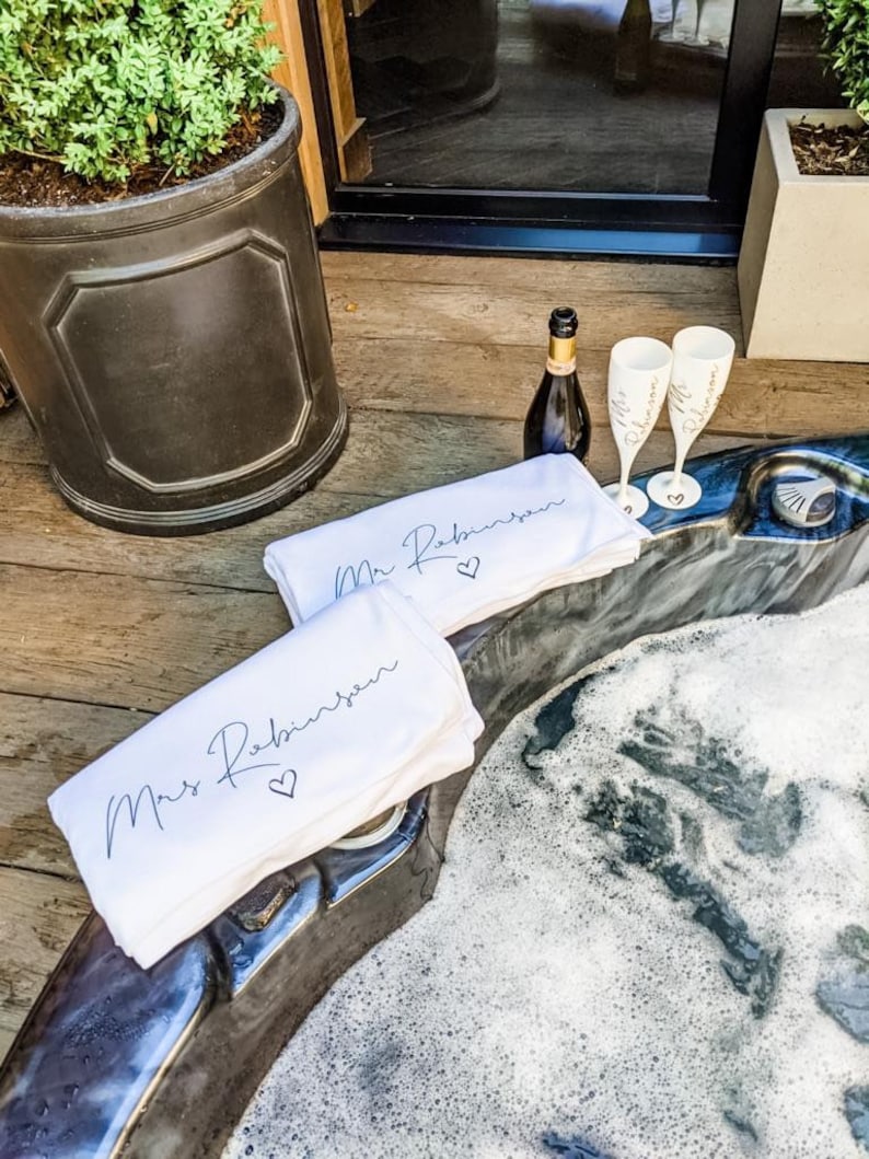 May include: Two white towels with the text "Mr. Robinson" and "Mrs. Robinson" with a heart symbol on each towel. The towels are placed on the edge of a hot tub with bubbles in the water. Two champagne flutes with the text "Mr. Robinson" and "Mrs. Robinson" are on the edge of the hot tub. A bottle of champagne is behind the flutes.