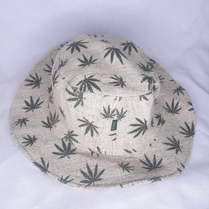 May include: A beige bucket hat with a green marijuana leaf pattern. The hat is made of a woven fabric.