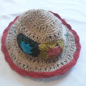 May include: A beige crocheted hat with a wide brim, featuring a red trim and a decorative floral accent. The hat has an open weave design. The floral accent includes blue, green, and pink elements.