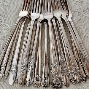 May include: A collection of antique silver forks, each with a unique handle design. The forks are arranged in a fan shape, showcasing the variety of floral and ornate patterns. The silver utensils are displayed on a white lace surface.