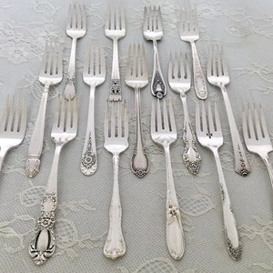 May include: An assortment of antique silver forks with various handle designs. The forks are arranged on a light blue lace tablecloth. The handles feature floral, geometric, and ornate patterns. The tines are evenly spaced and polished.