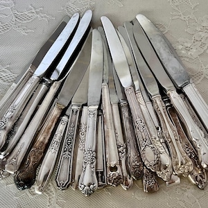 May include: A collection of vintage silver flatware knives with ornate handles. The knives are arranged in a fan shape, showcasing their intricate details.