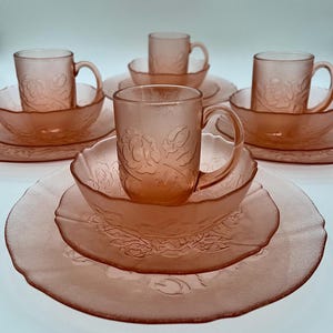 May include: A set of pink frosted glass dinnerware featuring a raised floral pattern. The collection includes multiple plates, bowls, and mugs with matching handles. Each piece displays a textured surface with embossed rose motifs throughout the set.