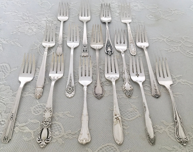 May include: An assortment of antique silver forks with various handle designs. The forks are arranged on a light blue lace tablecloth. The handles feature floral, geometric, and ornate patterns. The tines are evenly spaced and polished.