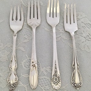 May include: Four silver forks with different handle designs are arranged on a white lace surface. The forks have ornate floral and scroll patterns. The tines are visible, and the forks are likely for a formal dining setting.
