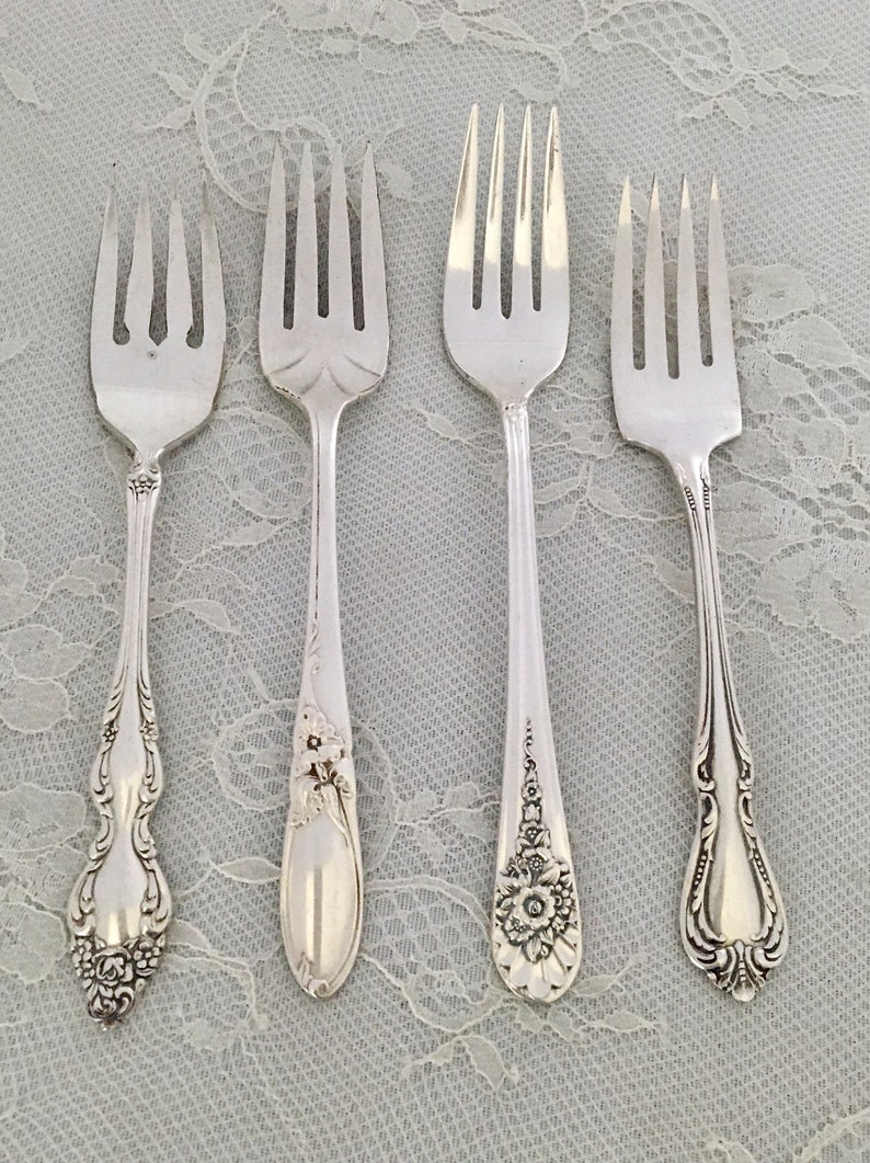 May include: Four silver forks with different handle designs are arranged on a white lace surface. The forks have ornate floral and scroll patterns. The tines are visible, and the forks are likely for a formal dining setting.