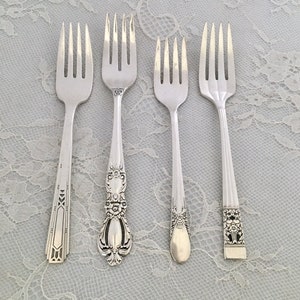 May include: Four silver forks with different handle designs are displayed. The forks have tines and varying handle patterns, including geometric, floral, and ornate details. The forks are arranged on a light-colored, lace-textured surface.