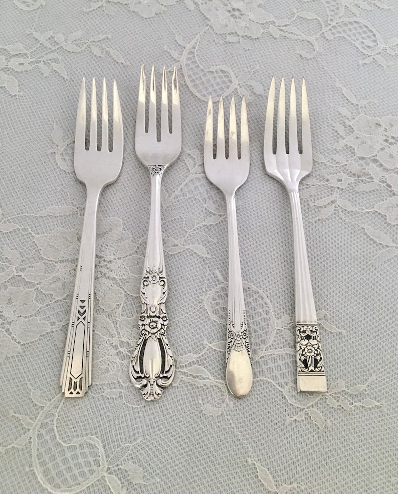 May include: Four silver forks with different handle designs are displayed. The forks have tines and varying handle patterns, including geometric, floral, and ornate details. The forks are arranged on a light-colored, lace-textured surface.