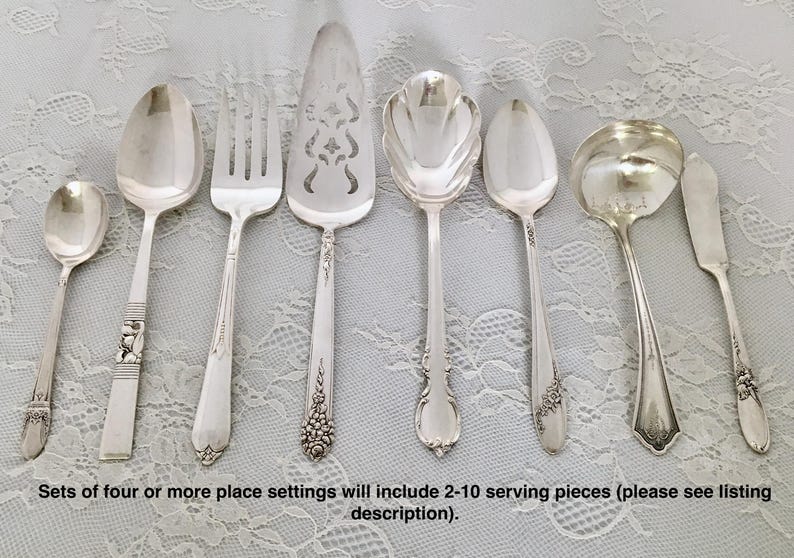 May include: A set of eight silver-plated serving pieces, including a serving spoon, a cake server, a butter knife, a gravy ladle, a sugar spoon, a pie server, a dinner fork, and a dinner spoon. The pieces are arranged on a white lace tablecloth.