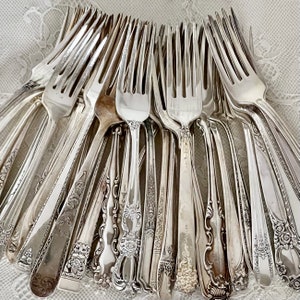 May include: A collection of antique silver forks, showcasing various designs and patterns. The forks are arranged in a circular fashion, with tines facing outwards. The silver cutlery reflects light, highlighting the intricate details of each piece.