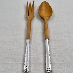 May include: A wooden fork and spoon set featuring light brown wooden heads and polished silver-tone metal handles. The handles are decorated with a beaded edge and a small, ornate floral-inspired motif at the base of each utensil.