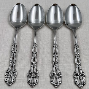 May include: Four identical silver-toned metal spoons arranged in a row. Each spoon features a polished oval bowl and a decorative handle with a series of open, teardrop-shaped cutouts and intricate scrollwork detailing at the base of the handle.