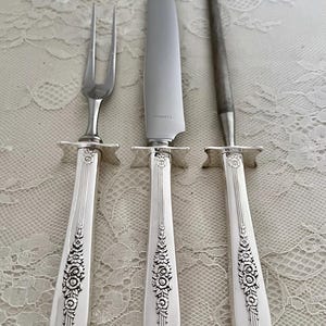 May include: A silver carving set featuring a fork, knife, and sharpening steel. The handles have a floral design and a decorative bolster. The knife blade is marked "STAINLESS". The set is displayed on a white lace tablecloth.