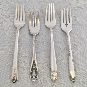 May include: Four silver forks with different handle designs are displayed on a white lace tablecloth. The forks vary in style, with ornate floral and geometric patterns on the handles. The tines are uniform, and the forks are arranged side by side.