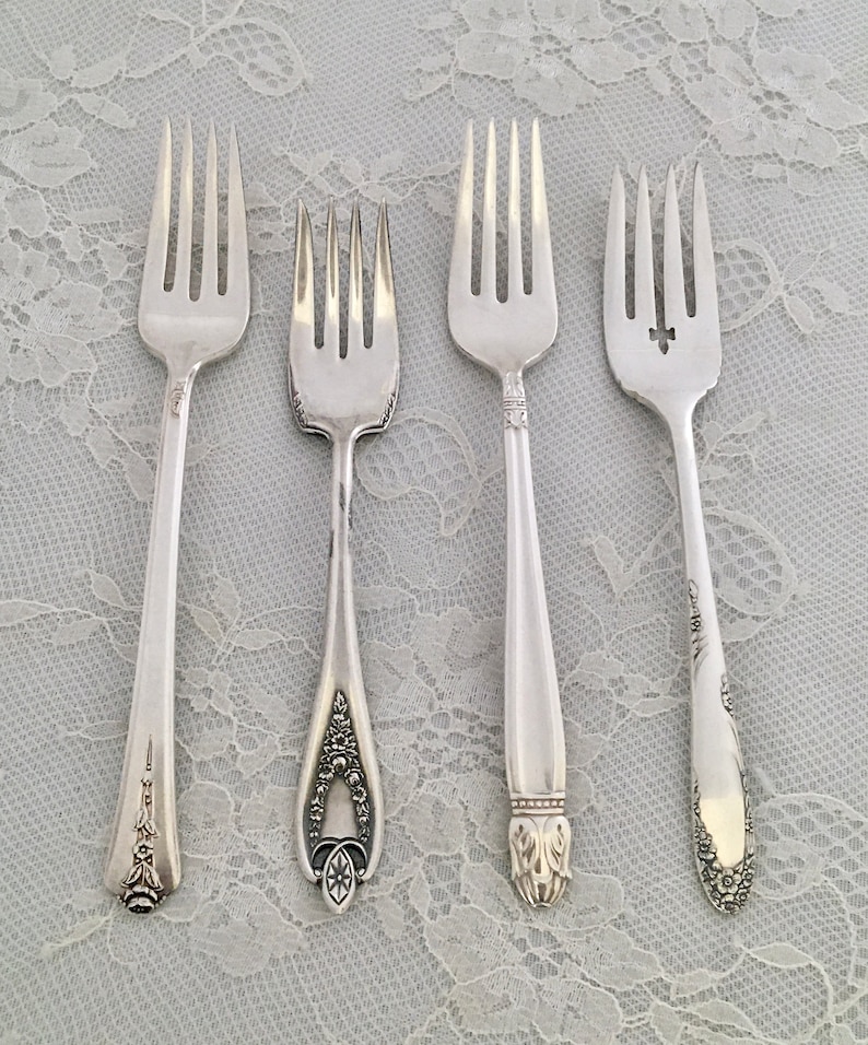 May include: Four silver forks with different handle designs are displayed on a white lace tablecloth. The forks vary in style, with ornate floral and geometric patterns on the handles. The tines are uniform, and the forks are arranged side by side.