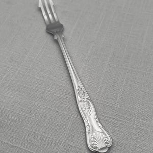 May include: A silver-toned metal cocktail fork featuring a three-tined head and a slender handle. The handle is decorated with an embossed floral motif near the center and a prominent shell-shaped design at the rounded base of the utensil.
