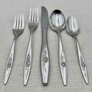 May include: A collection of five silver-coloured cutlery items: a fork, a knife, a spoon, and two additional forks and spoons. Each piece has a floral pattern on the handle. The utensils are displayed on a light-coloured, textured surface.