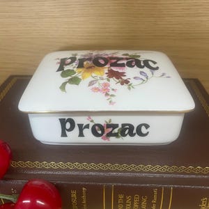 May include: A white ceramic trinket box with the word "Prozac" in black lettering on the lid and base. The lid is decorated with a floral design in yellow, pink, and burgundy. The box has a gold trim.