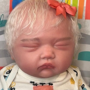 May include: A realistic baby doll with pale skin and short, wavy blonde hair. The doll has closed eyes, light eyebrows, and a small coral-colored ribbon bow attached to the hair. It wears a white short-sleeved bodysuit featuring a colorful pattern.