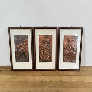 May include: Three rectangular wall art pieces in dark brown wooden frames with glass covers. Each frame contains a copper-toned metal relief plaque depicting scenes with figures, including a sailboat, a person holding a child, and a person with a tray.