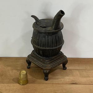 May include: A small, antique-style metal stove with a spout and a hinged lid. The stove is dark brown and sits on a decorative base. A gold thimble is next to the stove. The stove is likely a decorative item.