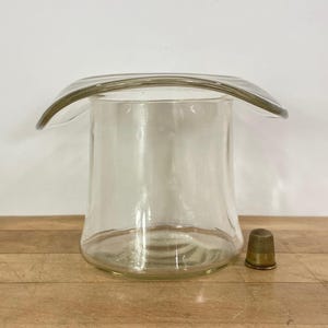 May include: A clear glass vessel shaped like an inverted top hat with a wide, curved brim and a cylindrical base. Beside the glass vessel sits a small, metallic, textured thimble with a gold-toned finish, providing a sense of scale.