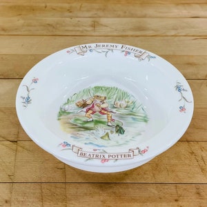 May include: White ceramic bowl featuring a colorful illustration of a frog fishing. The bowl has the text "MR JEREMY FISHER" and "BEATRIX POTTER" in banners. Floral accents decorate the rim, adding a touch of whimsy to this collectible item.