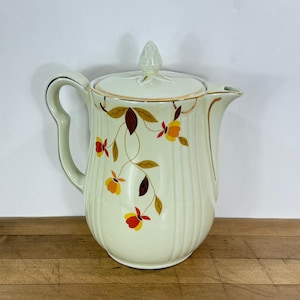 May include: Cream-colored vintage ceramic pitcher with lid. The pitcher features a floral design in yellow, orange, and brown. Gold trim accents the handle, spout, and lid. The pitcher has vertical ribbed detailing.