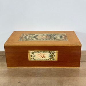 May include: A rectangular wooden box with a hinged lid, featuring a light brown finish. The top and front panels are decorated with rectangular inlays displaying intricate floral and scrollwork patterns in muted green, gold, and cream tones.