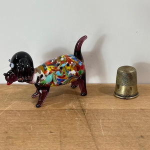 May include: A small glass dog figurine with a black head and a multicolored body featuring swirls of red, blue, yellow, and white. The dog has a dark purple tail and legs. Beside it sits a metallic, dimpled sewing thimble for scale.