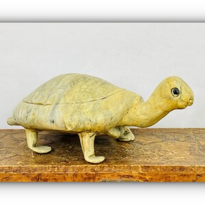 May include: A light yellow turtle-shaped decorative box. The turtle has a textured shell and a detailed head with black eyes. The box is resting on a wooden surface, and the background is a plain white wall.