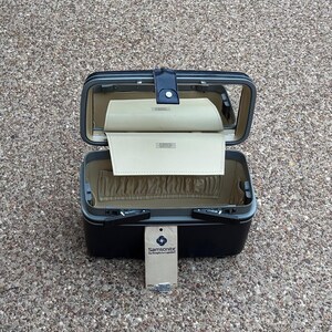 May include: A black Samsonite hard-shell vanity case with an open lid featuring an interior mirror and a beige fabric lining. The case includes a beige storage flap and an elasticized pocket. A rectangular Samsonite brand tag hangs from the front.