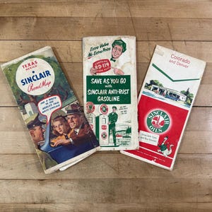 May include: Three vintage Sinclair paper road maps featuring colorful graphic illustrations. The left map shows Texas and Mexico, the center map promotes anti-rust gasoline with a cartoon mascot, and the right map displays Colorado and Denver.