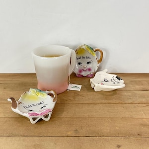 May include: A white and pink ombre mug with tea, a ceramic teapot-shaped tea bag holder, a face-shaped teacup, and a tea bag holder with the text "I will hold the bag". The items are on a wooden surface.