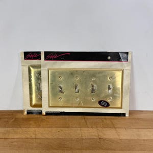 May include: Two gold-toned metal wall plates for four toggle switches, packaged in original cardboard backing with the brand name Style printed at the top. Each plate features a rectangular design with four cutouts for switches and mounting screws.