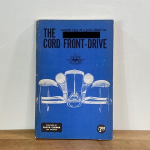 May include: A vintage book titled "The Cord Front-Drive" with a blue cover. The cover features a white illustration of a classic car and the text "complete story of a great classic car". Published by Floyd Clymer, Los Angeles.