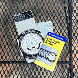 May include: A collection of aviation navigation tools, including a flight computer, a slide graphic computer manual, and a map. The flight computer is circular with various scales and markings. The manual is yellow and blue, with the text "Slide Graphic Computer Manual."