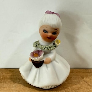 May include: A ceramic figurine of a person with white hair styled in a bun with a purple bow. The figure wears a white dress and a green textured lei around the neck. They are holding a brown basket containing a pink and yellow flower.
