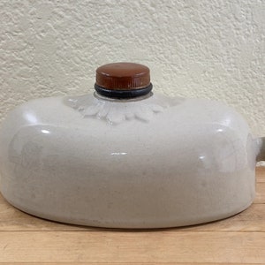 May include: A cream-colored, ceramic hot water bottle with a brown stopper and handle. The bottle has a rounded shape with a decorative floral design near the stopper. The handle is also brown.