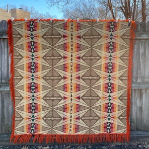 May include: A beige and brown patterned blanket with geometric designs and vertical stripes of orange, pink, and green. The blanket has orange fringe along the edges and is hanging on a wooden fence.