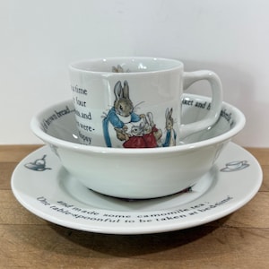 May include: A white ceramic tea set featuring illustrations of rabbits and text. The set includes a mug, a bowl, and a plate. The plate and bowl have text around the edges. The mug is stacked on the bowl, which is on the plate.
