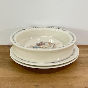 May include: Cream-colored ceramic bowl and two plates. The bowl has a wide rim with text around the edge and a detailed illustration in the center. The plates are stacked beneath the bowl, also with text around the edges.