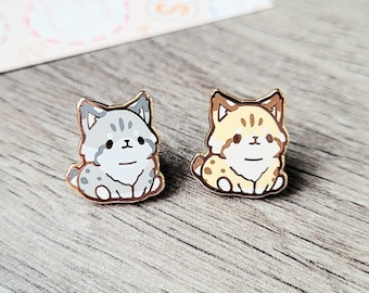 May include: Two enamel pins featuring cartoon bobcats. The bobcat on the left is gray and white, while the bobcat on the right is yellow and white.
