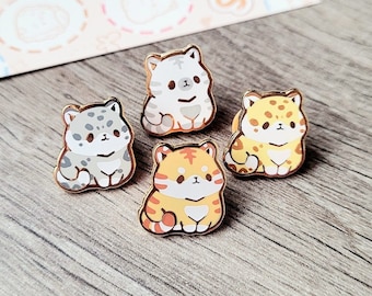 May include: Four enamel pins featuring different cartoon cats. The cats are all sitting down and have different colored fur. The pins are gold-colored and have a shiny finish.