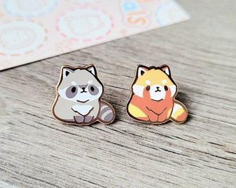 May include: Two enamel pins featuring cartoon animals. The first pin is a gray raccoon with a white belly and black eyes. The second pin is a red panda with a white belly and black eyes. Both pins have gold metal backing.