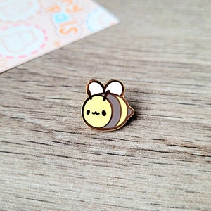 May include: A gold-coloured enamel pin featuring a cartoon bee with a yellow body, black stripes, and a cute face. The bee has small white wings and is facing to the left.