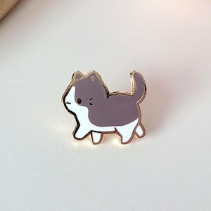 May include: A gold-colored enamel pin in the shape of a cartoon cat. The cat is depicted in a walking pose, with a white belly and face, and a gray back and head. The pin is a cute accessory.
