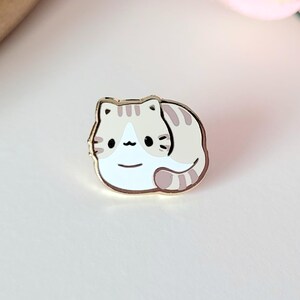 Könnte beinhalten: Ein goldfarbener Emaille-Pin mit einer Cartoon-Katze, die sich zusammengerollt hat. Die Katze hat ein weißes und graues Streifenmuster und ein lächelndes Gesicht. Der Pin befindet sich auf einer weißen Oberfläche, mit einem Holztablett und einer rosa Rose im Hintergrund.
