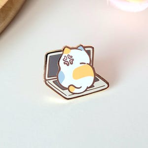 May include: A gold-colored enamel pin featuring a cartoon cat sitting on a laptop. The cat has white, blue, and yellow fur and an angry expression. The laptop screen is gray.